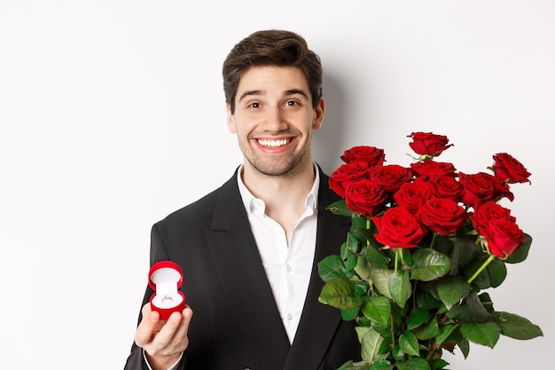 Primo piano di un uomo attraente in completo, con in mano un mazzo di rose e un anello di fidanzamento, che fa una proposta, in piedi su uno sfondo bianco