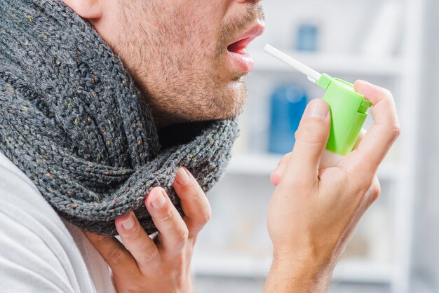 Primo piano di un uomo che indossa una sciarpa grigia intorno al collo usando uno spray per trattare il mal di gola