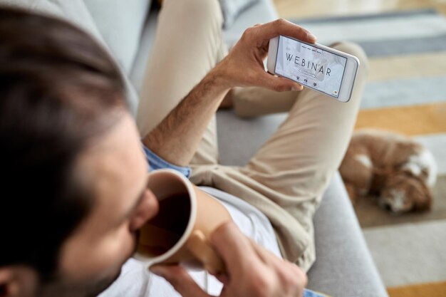 Primo piano di un uomo che usa lo smartphone e segue il webinar mentre beve caffè a casa