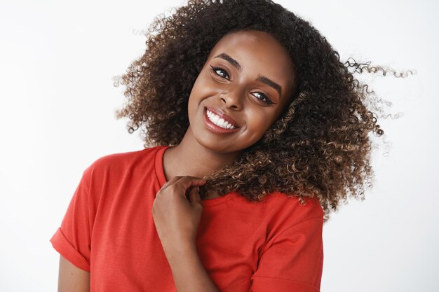 Primo piano di una bella donna afroamericana positiva energica e attiva che si sveglia felicemente iniziando la giornata con il jogging indossando una t-shirt rossa inclinando la testa ottimista e adorabile sul muro bianco