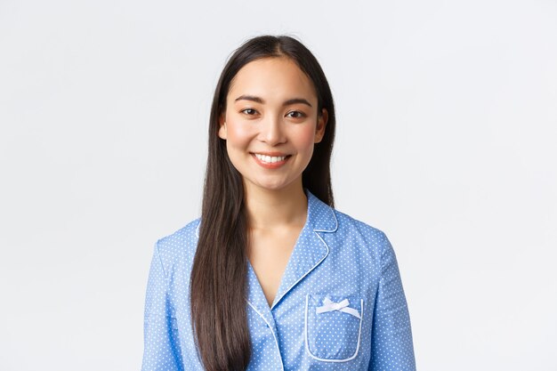 Primo piano di una bella ragazza asiatica in pigiama blu con denti bianchi perfetti, sorridendo alla telecamera felice, svegliandosi al mattino entusiasta, dormendo bene la notte, in piedi sfondo bianco.