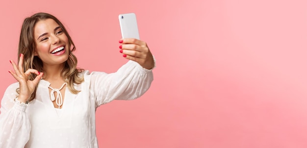 Primo piano di una bella ragazza bionda soddisfatta in abito bianco che prende selfie registra video sul telefono cellulare