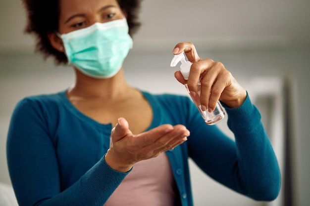 Primo piano di una donna afroamericana che indossa una maschera facciale mentre si disinfetta le mani a casa