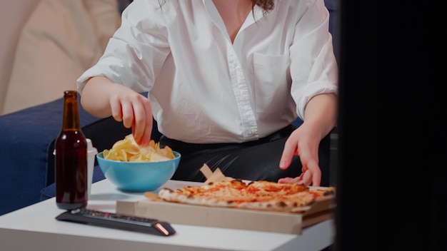 Primo piano di una donna che mette una scatola di pizza sul tavolo mentre beve bevande e una ciotola di patatine. Persona caucasica che si prepara a mangiare un pasto da asporto da un fast food in soggiorno