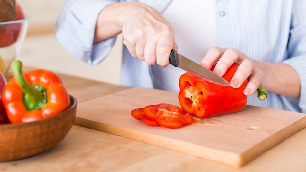 Primo piano di una donna che taglia il peperone dolce rosso fresco con il coltello sul tagliere di legno