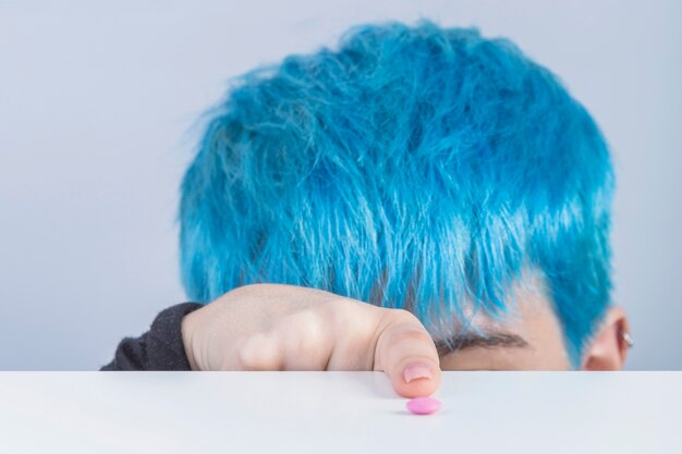 Primo piano di una donna con capelli tinti di blu che punta il dito verso la pillola rosa sul tavolo