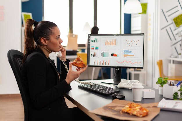 Primo piano di una donna d'affari seduta alla scrivania davanti al computer che mangia una fetta di pizza