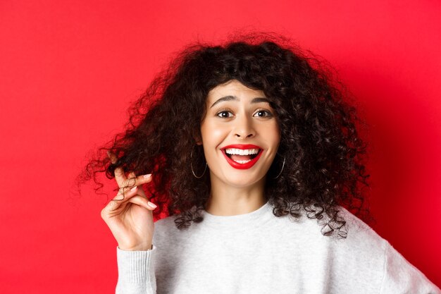 Primo piano di una donna felice che gioca con i capelli ricci e ride di qualcosa di divertente in piedi su sfondo rosso...