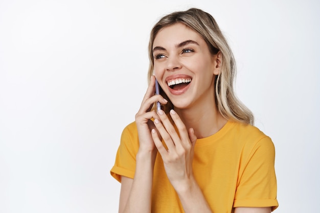 Primo piano di una donna felice che parla al cellulare, ride su chiamata, in piedi su bianco in maglietta gialla.