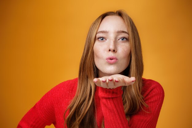 Primo piano di una donna rossa sensuale e attraente in abito di maglia rosso piegando le labbra in avanti mentre invia un bacio d'aria alla telecamera dando mwah che tiene il palmo vicino alla bocca sul muro arancione