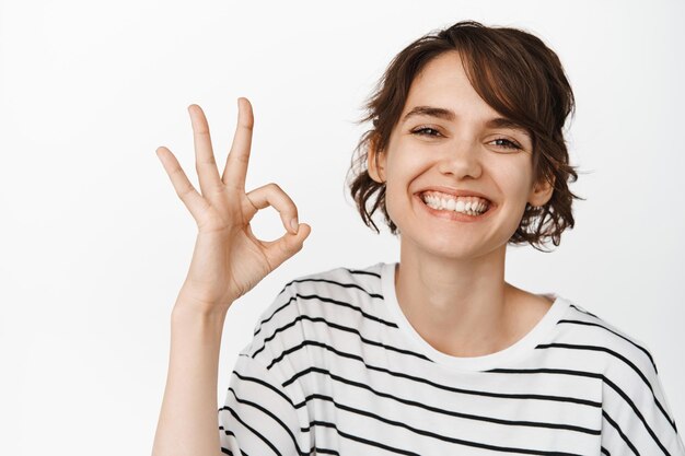 Primo piano di una donna sorridente felice che mostra ok, ok firmare per l'approvazione, mi piace e sono d'accordo, elogiare l'eccellente scelta, in piedi in maglietta su sfondo bianco.