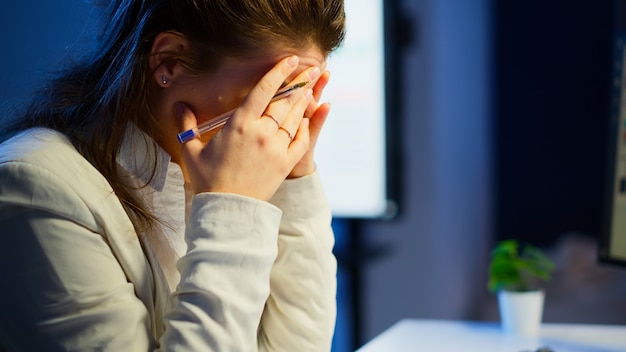 Primo piano di una donna stressata e impegnata con mal di testa che lavora dall'ufficio a tarda notte. Dipendente stanco e concentrato che utilizza la moderna tecnologia di rete wireless che fa gli straordinari per i rapporti di controllo del lavoro