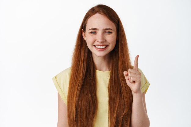 Primo piano di una giovane donna allegra con i capelli rossi e un sorriso bianco, che punta il dito in piedi in maglietta su bianco.