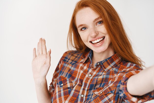 Primo piano di una giovane donna con lunghi capelli rossi e occhi azzurri che prende selfie e dice ciao Ragazza blogger agitando la mano e sorridendo alla telecamera