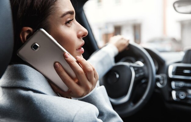 Primo piano di una giovane donna d'affari che guida in ufficio e chiama qualcuno al telefono Donna seduta sul conducente sedersi e parlare sullo smartphone