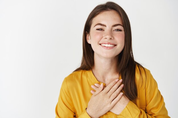 Primo piano di una giovane donna felice e premurosa che guarda grata davanti, sorride mentre si tiene per mano sul cuore, dice grazie, essendo grata, in piedi sul muro bianco