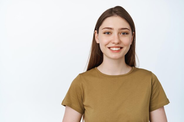 Primo piano di una giovane donna felice, sorridente con i denti bianchi, procedura di sbiancamento presso la clinica dentale, in piedi in maglietta su sfondo bianco.