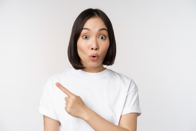 Primo piano di una giovane donna giapponese sorridente che punta il dito a sinistra nello spazio della copia che mostra l'annuncio o il banner pubblicitario in piedi su sfondo bianco