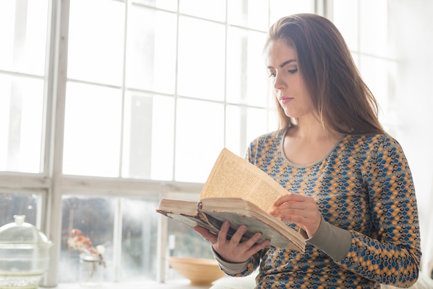 Primo piano di una giovane donna in piedi vicino al libro di lettura della finestra