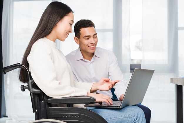 Primo piano di una giovane donna sorridente che si siede sulla sedia a rotelle che mostra al suo collega sul computer portatile