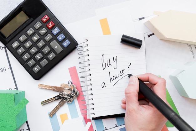 Primo piano di una persona che scrive per comprare una casa sul blocco note a spirale con le chiavi; calcolatrice e modello di casa