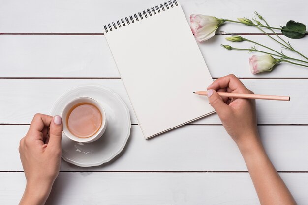Primo piano di una persona che scrive sul blocco note con la tazza di tettarella sulla scrivania in legno