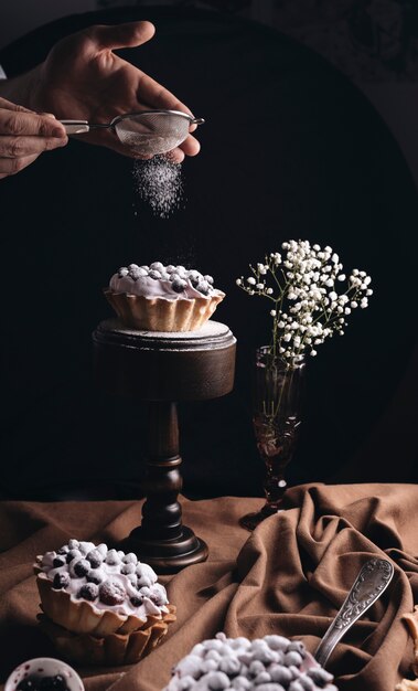 Primo piano di una persona che spolvera la polvere dello zucchero sulla torta di frutta con il vaso di fiori del respiro del bambino