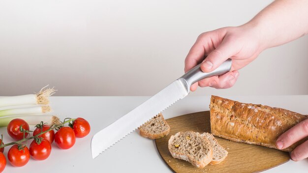 Primo piano di una persona che taglia pane con il coltello affilato sullo scrittorio bianco contro fondo bianco