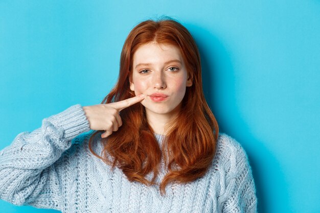 Primo piano di una ragazza adolescente sfacciata con i capelli rossi, che indica la sua guancia e fissa la telecamera, in piedi su sfondo blu.