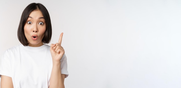 Primo piano di una ragazza asiatica creativa che alza il dito suggerendo che smth ha un'idea rivolta verso l'alto segno eureka in piedi su sfondo bianco