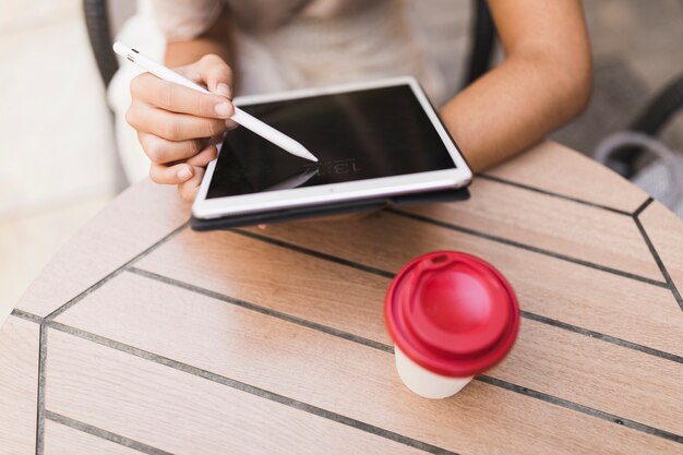 Primo piano di una ragazza che utilizza lo stilo sulla compressa digitale con la tazza di caffè asportabile sulla tavola