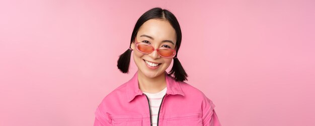 Primo piano di una ragazza coreana alla moda in occhiali da sole che sorride felice in posa su sfondo rosa Concetto di faccia della gente