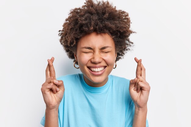 Primo piano di una studentessa diligente felice dai capelli ricci esprime un desiderio prima che l'esame attenda i sogni che si avverano incrocia le dita sorride positivamente indossa abiti blu casuali isolati sul muro bianco