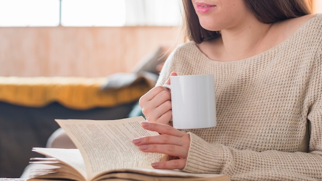 Primo piano di una tazza di caffè della holding della giovane donna che gira la pagina del libro