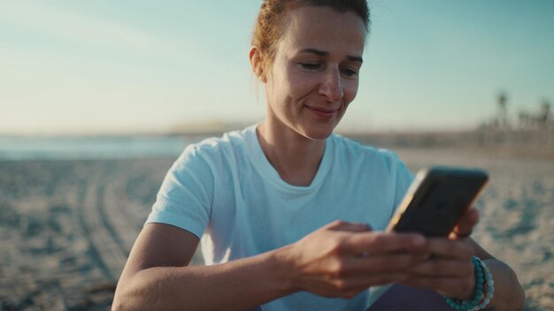 Primo piano donna che controlla i suoi social media utilizzando lo smartphone in riva al mare Ragazza sportiva che riposa dopo l'allenamento sulla spiaggia