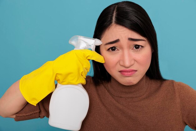 Primo piano esausto stressato cameriera asiatica oberata di lavoro dai lavori domestici con guanti gialli che tengono uno spray vicino al suo tempio, concetto di casa di pulizia, donna triste e infelice spaventata sopraffatta