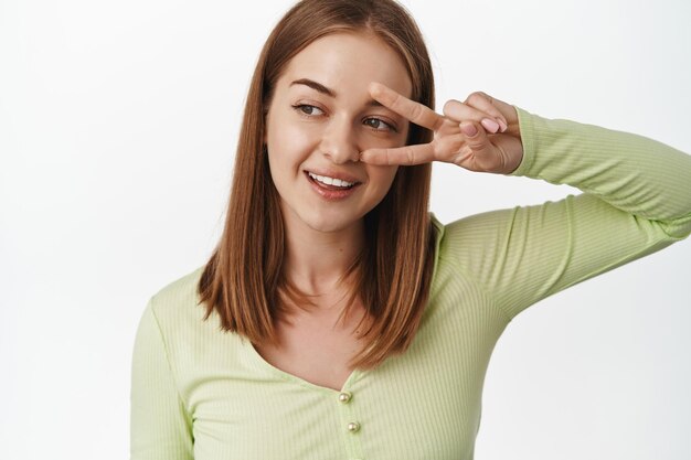 Primo piano giovane donna bionda in verde elegante bloud, mostra il gesto del segno v di pace vicino all'occhio, sorridendo e guardando a sinistra al testo promozionale di bellezza, in piedi su sfondo bianco.