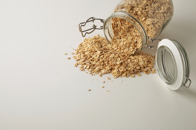 Primo piano in pieno vaso rustico aperto con fiocchi d'avena sani sparsi isolati nel lato sulla vista laterale del tavolo bianco