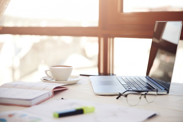 Primo piano laterale di una scrivania con computer portatile, tazza di caffè, occhiali e articoli di cancelleria