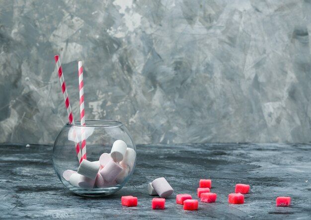 Primo piano marshmallow e canne da zucchero in vaso con caramelle e tovaglia a quadretti rossi su superficie di marmo blu e grigio scuro. spazio libero orizzontale per il testo