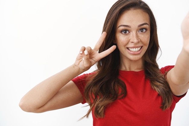 Primo piano ottimista tenero femminile bruna giovane donna tenere smartphone una mano che mostra buona volontà e benessere segno vittoria o gesto di pace sorridente sciocco esprimere entusiasmo sfondo bianco