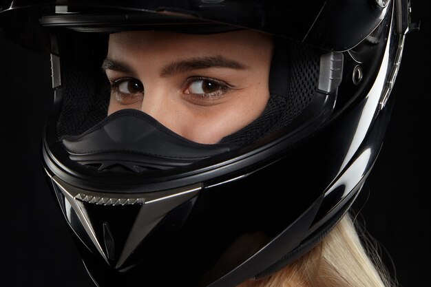 Primo piano ritratto di corridore motociclista femminile caucasico con occhi felici che indossa il casco di sicurezza moderno nero, andando alla concorrenza, sentendosi eccitato. Concetto di velocità, estremo, pericolo e attività