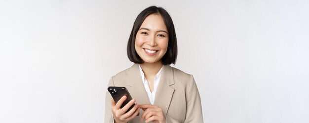 Primo piano ritratto di donna coreana donna aziendale in tuta utilizzando il telefono cellulare e sorridente tenendo lo smartphone in piedi su sfondo bianco