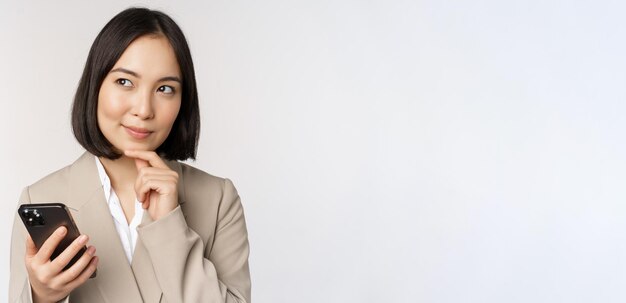 Primo piano ritratto di donna coreana donna aziendale in tuta utilizzando il telefono cellulare e sorridente tenendo lo smartphone in piedi su sfondo bianco