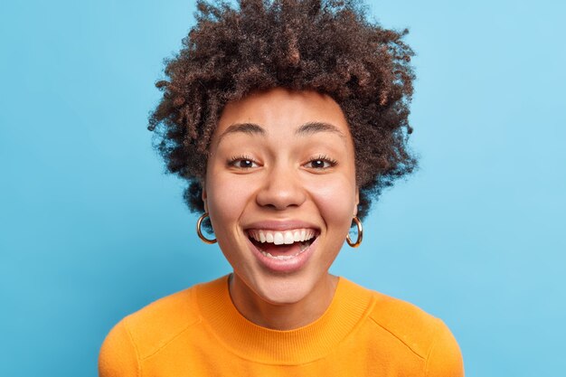 Primo piano ritratto di donna dalla pelle scura con capelli ricci naturali, pelle sana pulita, sorrisi, esprime ampiamente la felicità, ha denti bianchi perfetti, indossa abiti casual isolati sul muro blu.