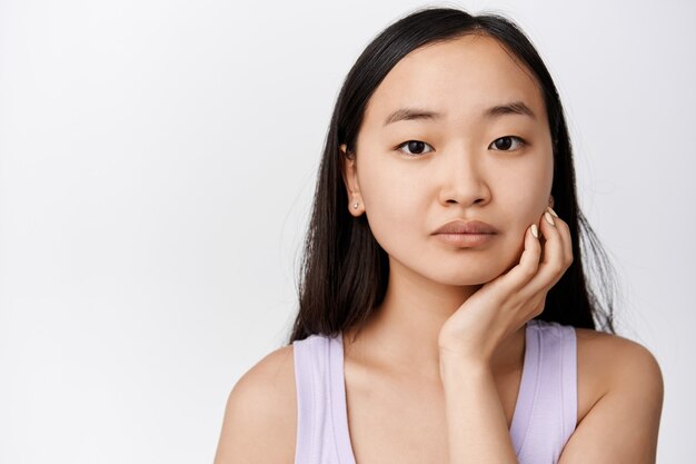 Primo piano ritratto di ragazza asiatica che tocca il viso e guarda il bianco in piedi sul bianco.