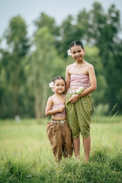 Primo piano, ritratto di sorella carina e giovane sorella in abito tradizionale tailandese e mettere un fiore bianco sul suo orecchio in piedi nel campo di riso, sorriso, concetto di amore tra fratelli, spazio di copia