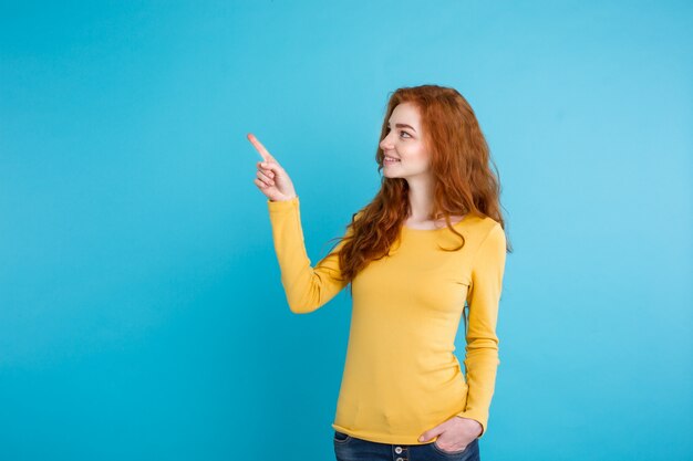Primo piano Ritratto giovane bella ragazza attraente redhair felice con qualcosa e dito di puntamento. Sfondo blu pastello. Copia spazio.