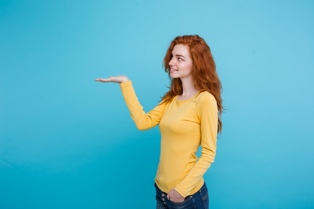 Primo piano Ritratto giovane bella ragazza attraente redhair felice con qualcosa e dito di puntamento. Sfondo blu pastello. Copia spazio.