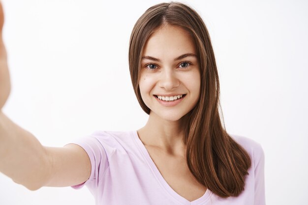 Primo piano sparato di affascinante giovane bruna femmina europea felice con capelli lunghi e forti e pulire sking sorridente amichevole mentre si tira la mano in avanti come se si stesse prendendo selfie